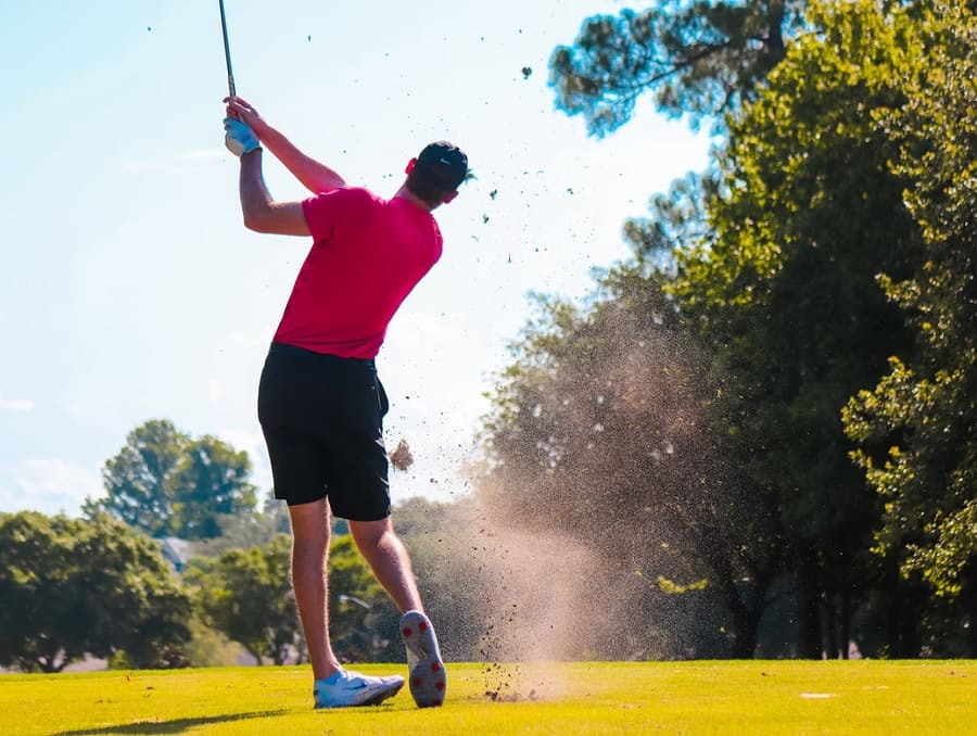 A golfer in a red shirt swings, hitting the turf and knocking dirt and grass into the air