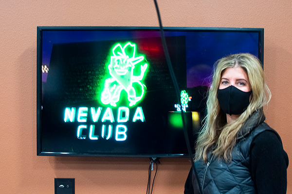 Janice Baker stands in front of a television screen with a neon green Nevada Club sign behind her.