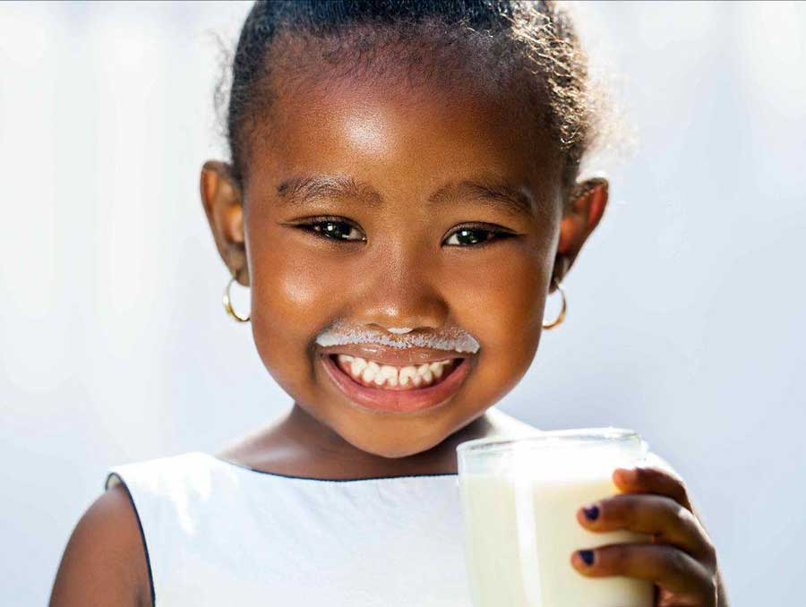 A child holding a glass of milk smiles with a big milk moustache on her face.