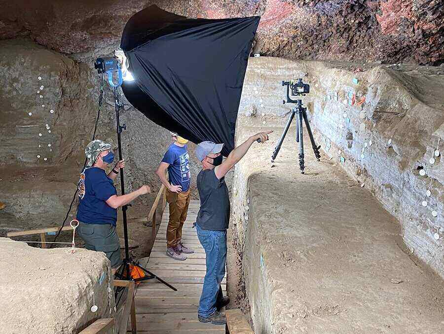 Three people stand in an underground cave observing a section of the cave’s interior wall that was part of an archaeological dig in 1979-80