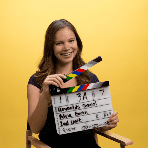 Adria Borich holds a clapboard in front of a yellow background.