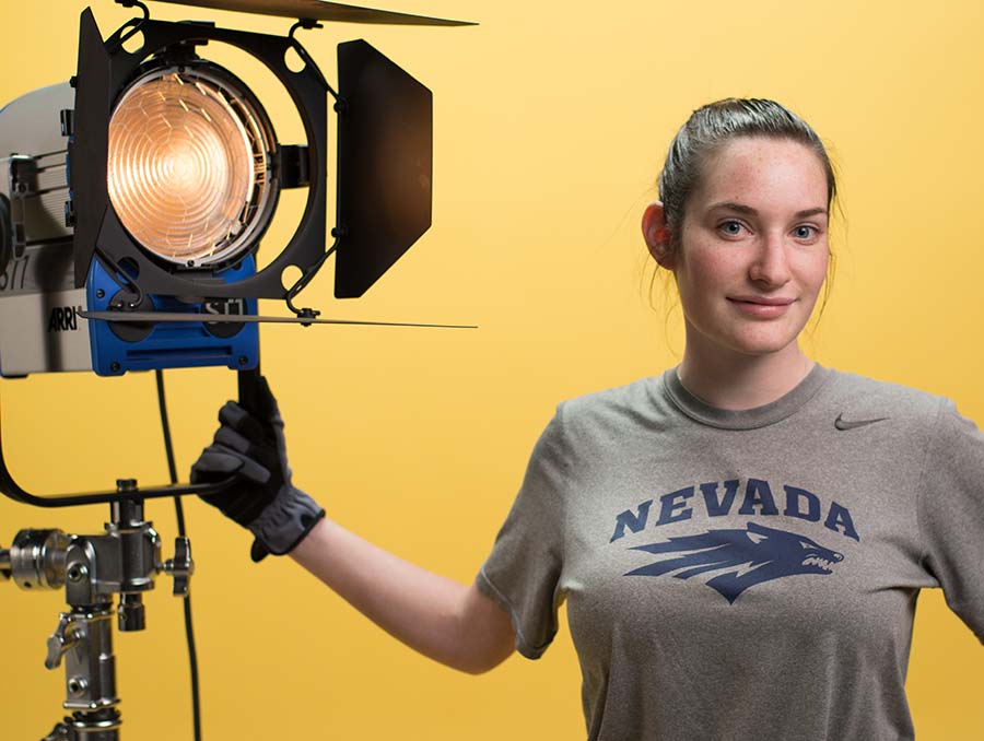 A woman wearing a University of Nevada, Reno t-shirt smiles to the camera while holding a light in front of a yellow background. 