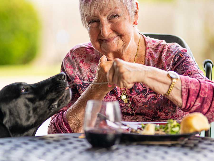 An older adult with pink hair smiles as she shares a bite of food from her meal with a big black dog.