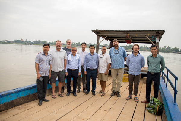 A group of men stand on a dock with lush foliage lining the river banks behind them.