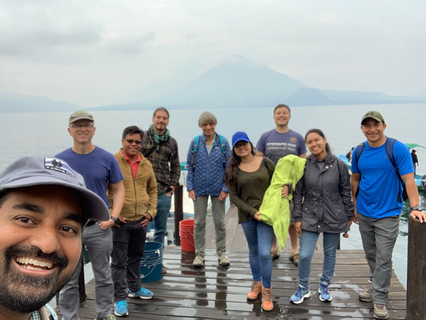 Chandra se toma una selfie con un grupo de personas en un muelle sobre un lago, con una gran montaña al fondo.