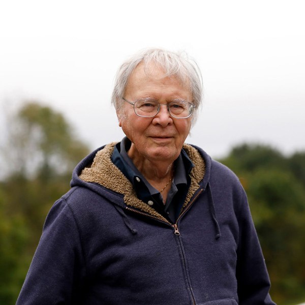 Wally Broecker smiles, wearing a blue polo and sweatshirt in front of green foliage in the background.