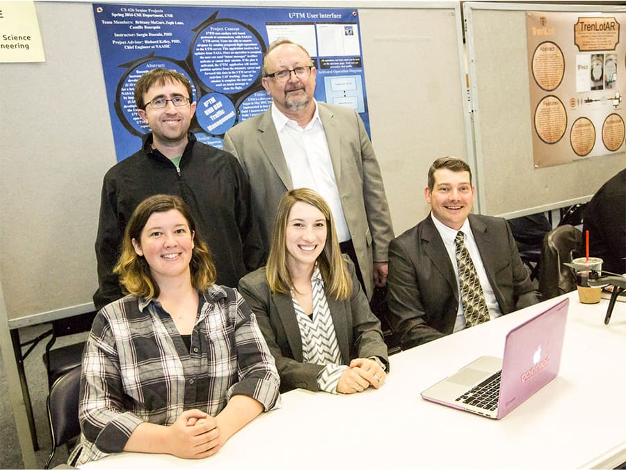 Two people standing and three sitting in front of a project poster with a table in front of them.