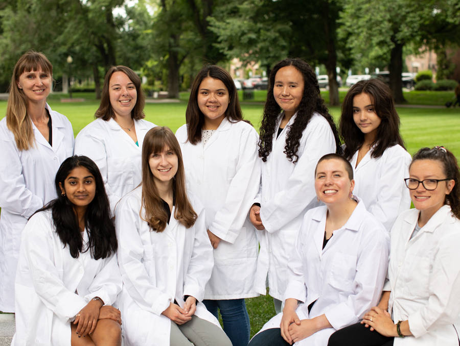 Nine women in white lab coats smile for a photo on the green University quad.