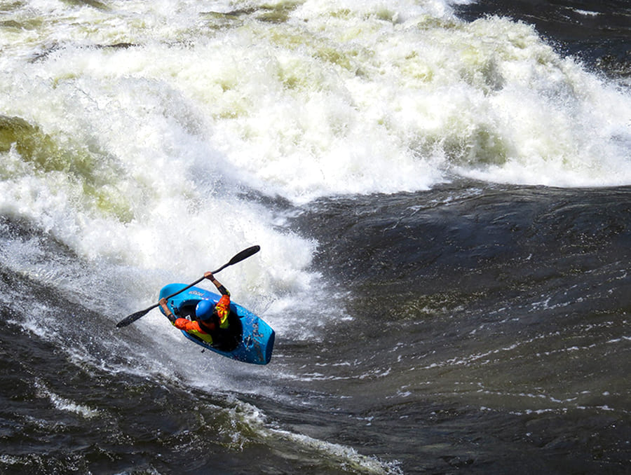 Brooke Hess kayaking in a blue kayak in a river near a white water rapid.