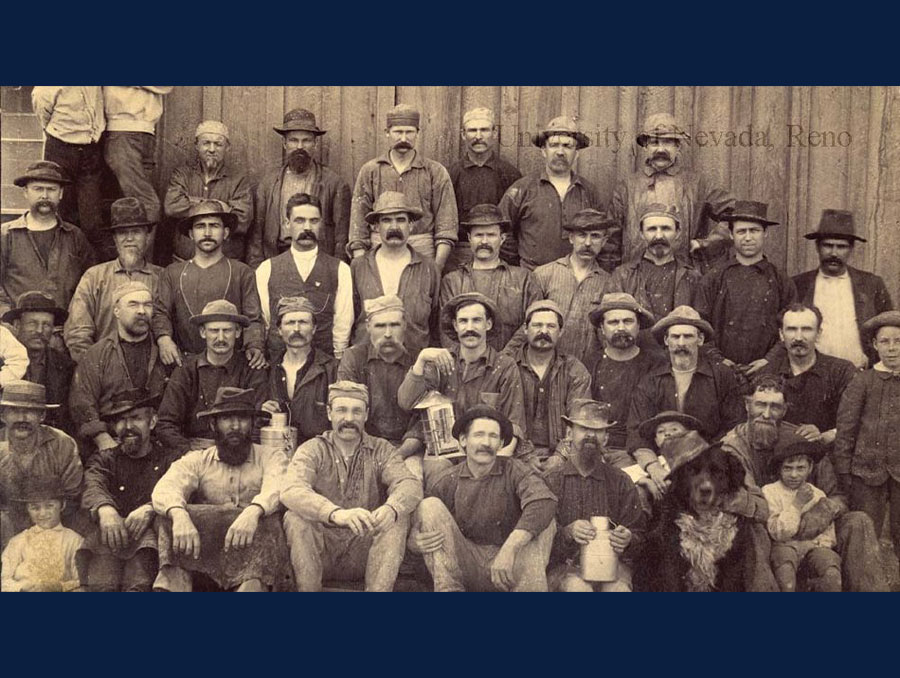 Historical photograph of a group of miners, children, and a black dog at the Gould and Curry mine in Virginia City, around 1888.