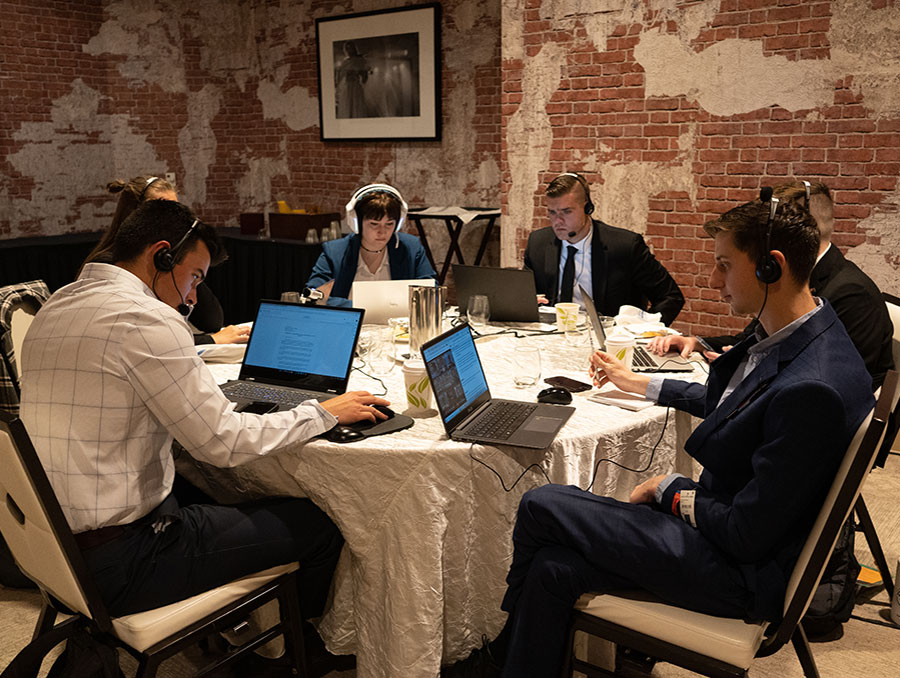 Students sit around a table inside with headsets on debating other students on computers