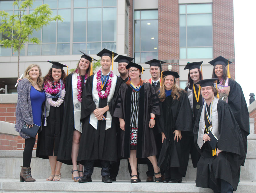 Eleven people stand, ten of whom are wearing graduation regalia, and smile on the steps outside of the Pennington Student Achievement Center.