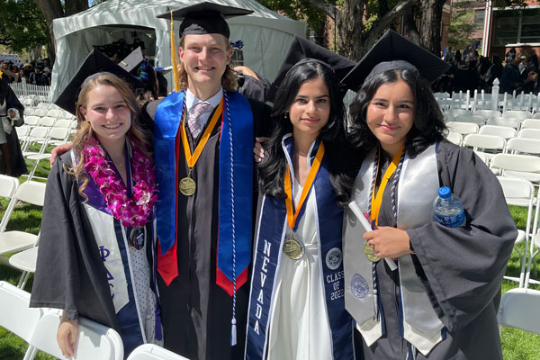 Four students wearing graduation regalia stand and smile on a sunny day. They are between rows of white chairs.