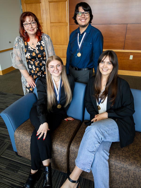 Two women sit in chairs. A woman and man stand behind them. The man and the women who are sitting are wearing medals.