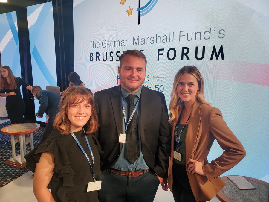 Three student attendees pose for a photo in front of the German Marshall's Fund Brussels Forum event sign.
