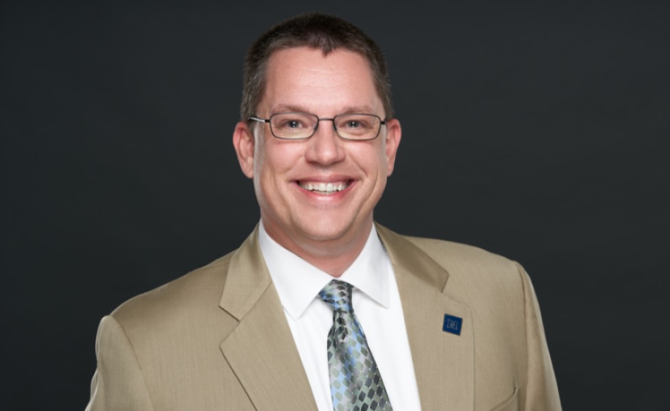 Dr. Frederick Steinmann poses in a tan suit in front of a dark gray background