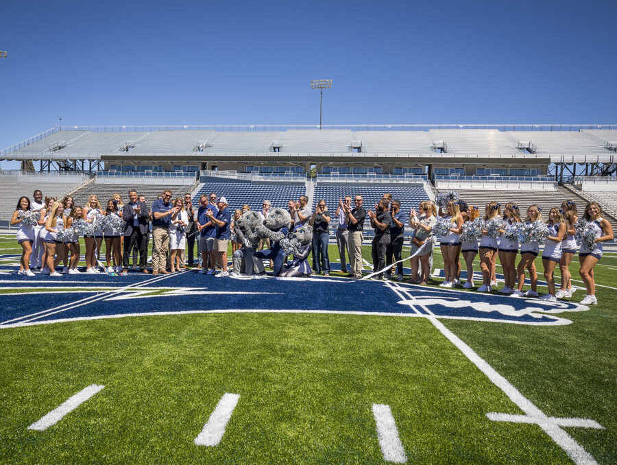 Ribbon cutting ceremony with Brian Sandoval, ITS Logistics leadership, and Wolf Pack Football leadership along with the Nevada Wolf Pack Cheerleaders and mascots on the new turf during a sunny day. Everyone is happy, smiling, and you can see the freshly cut ribbon hanging amongst the crowd of nearly 50 people. 
