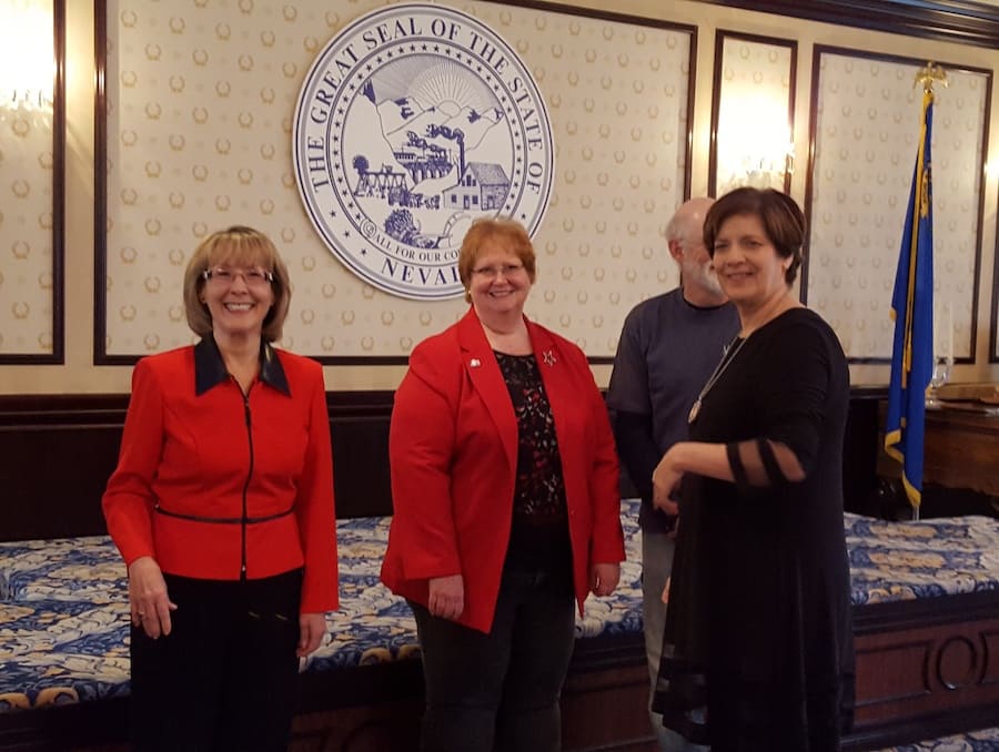 Three women, two in red and one in black, posing with the Seal of Nevada in the background.