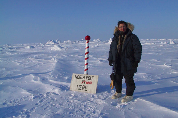A man, wearing heavy winter clothing, stands in a snowy landscape next to a red and white pole with a sign below that reads, "North Pole is here" with the word "is" crossed out and "was" written next to it.
