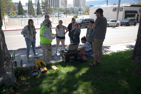 Students wearing gold lanyards stand around a man in a neon yellow vest as he explains the equipment next to him.