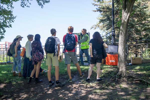 Students stand around a man in a neon yellow vest explaining the equipment he is using. They are standing next to some trees.