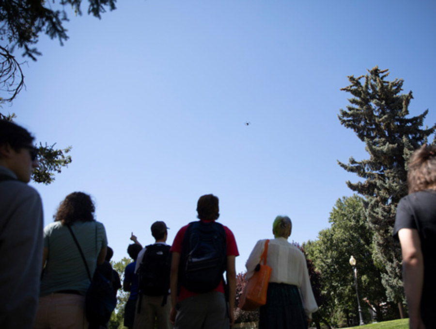 A group of students stands, looking upward at a drone against a clear blue sky. There are trees at the right and left edges of the photo.
