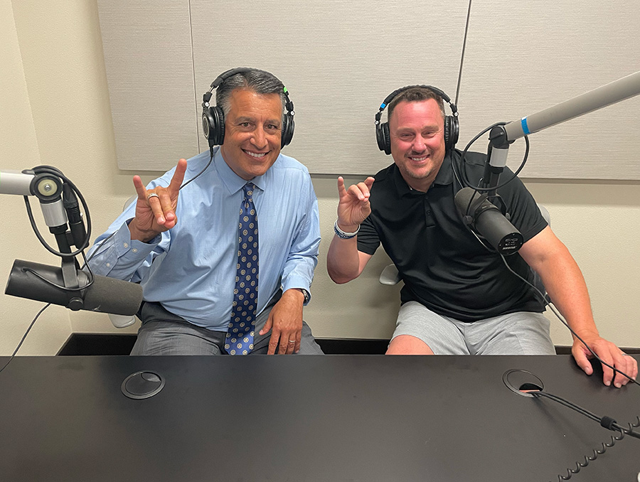 President Brian Sandoval and Coach Ken Wilson smile while sitting next to each other, wearing headphones in the recording space