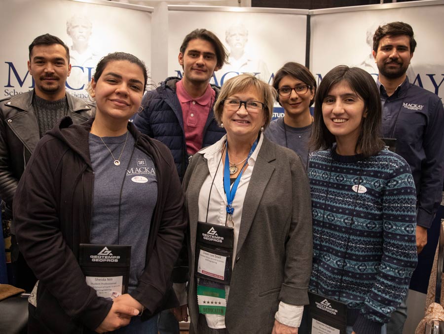 Seven people stand in front of three Mackay School posters. There are three people in the front row and four in the back, all are smiling.