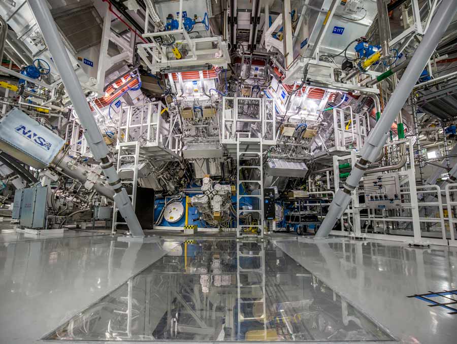 In a large room, a circular device three stories tall with wires and panels all over it sits with scaffolding around it to access various parts of the machine.