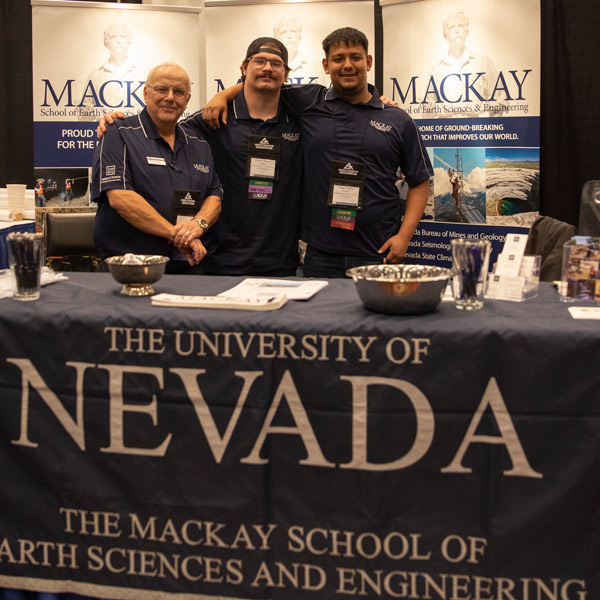 Three people stand in the Mackay School booth at the conference. They are all wearing Mackay polos and smiling with their arms around one another. There is Mackay swag on the table in front of them.