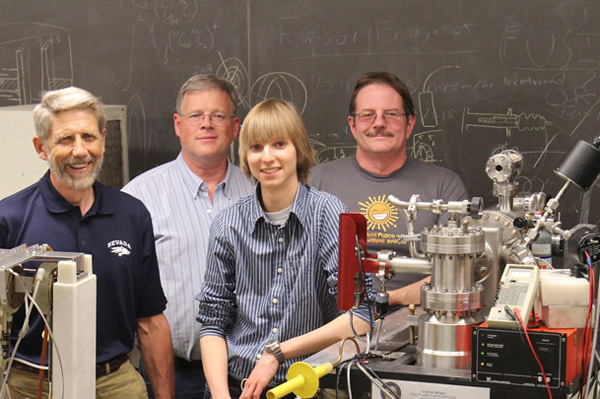 Four people stand in a row in front of a blackboard and behind a table with various machines on it. One of the people is a teenager.