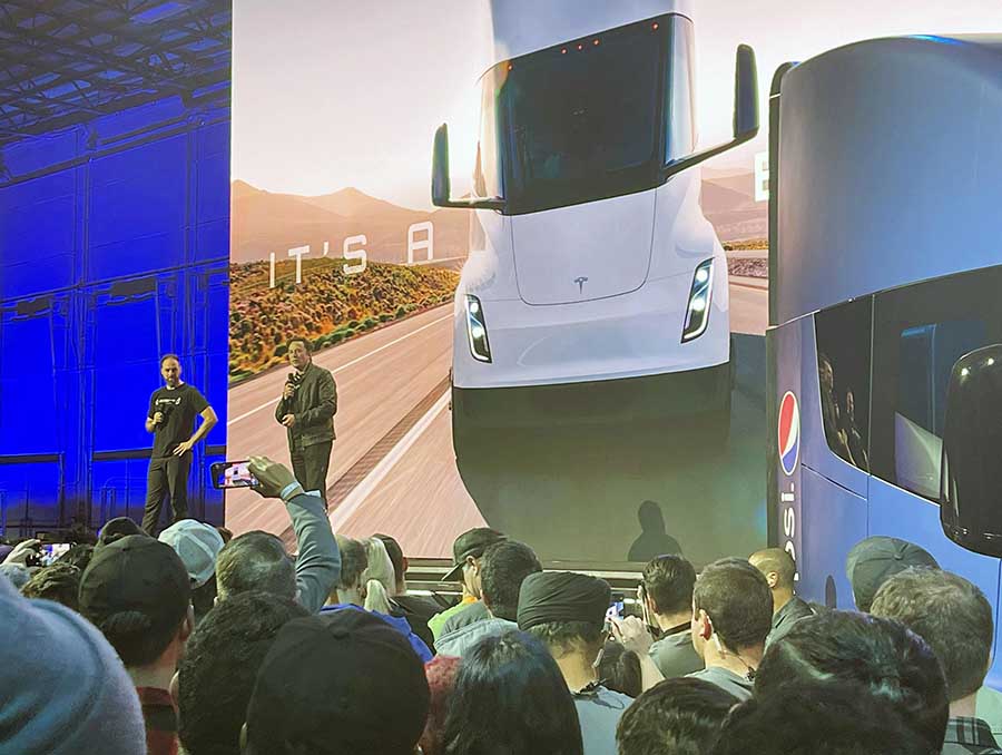 Tesla CEO Elon Musk and an unnamed man stand on a stage in front of a crowd, a large picture of a semi truck behind them.
