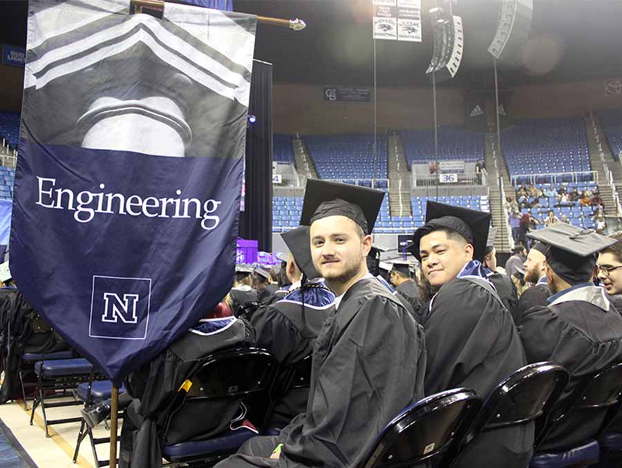 Two male students in cap and gown attire seated indoors next to a banner that reads "Engineering."