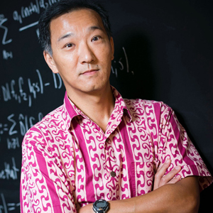 Ken Ono smiles in front of a blackboard with formulae written on it with his arms crossed, wearing a pink shirt.