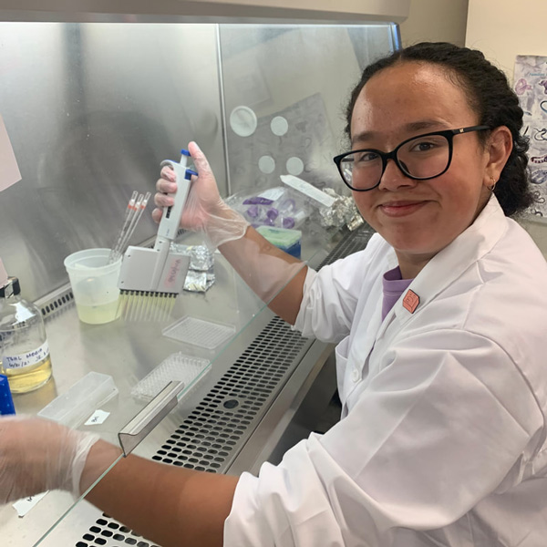 Jennifer Rodriguez smiles at the camera while working in a fume hood with a pipette in her hand and wearing a lab coat.