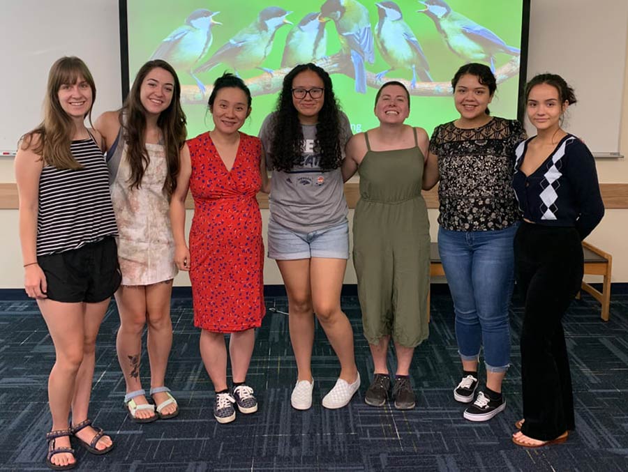 Seven women stand and smile in front of a screen with birds on it.