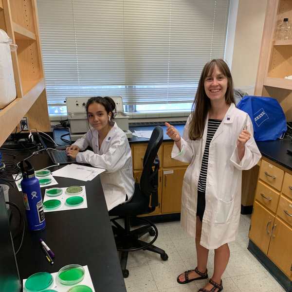 Ruby Pacheco sits at a computer next to green agar plates, with Jamie Voyles standing next to her and giving her two thumbs up.