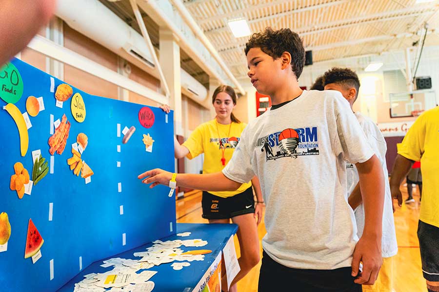 In a brightly lit gymnasium, kids wearing Hoops for Hope shirts arrange foods on a board by their healthiness, with healthy foods going in the 'Go' category, unhealthy foods going in the 'Whoa' category and in between foods going in the 'Slow' category.