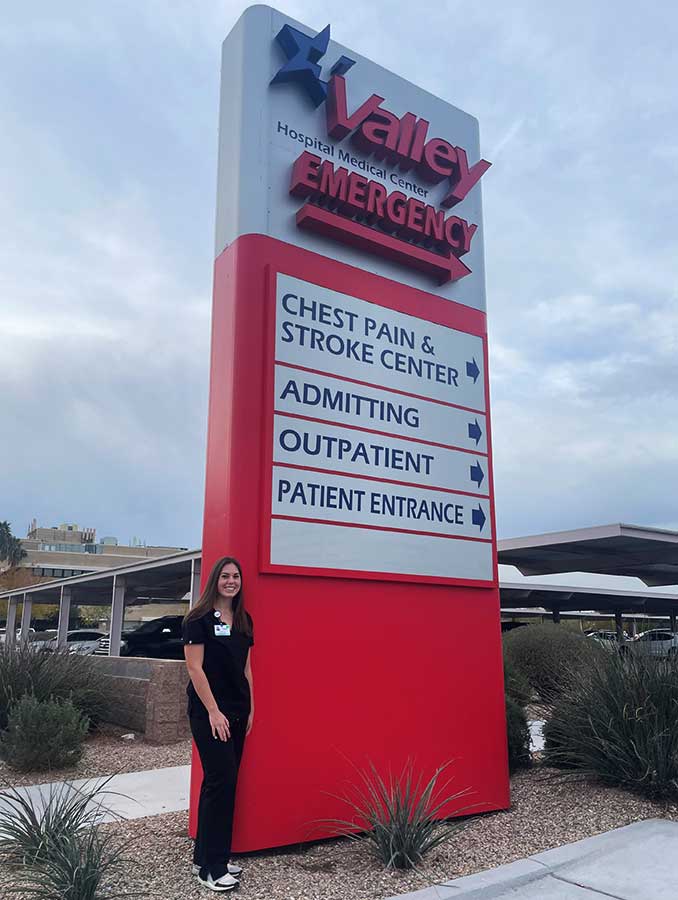 Samantha Reineck in black scrubs in front of Valley Medical Center's emergency sign that directs patients to the chest pain and stroke center, admitting, outpatient or patient entrance.