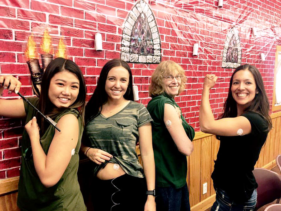 Four women show off their demo infusion sets in front of castle-themed decorations. One points to her set using a magic wand. Another flexes her biceps to show off her set. A third lifts her shirt to show where her set is attached to her stomach.