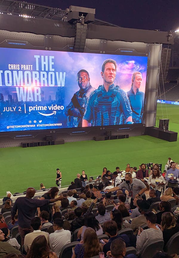 An audience sitting in chairs look toward a screen with the words "The Tomorrow War" and a picture of Sam Richardson, Chris Pratt and Yvonne Strahovski.