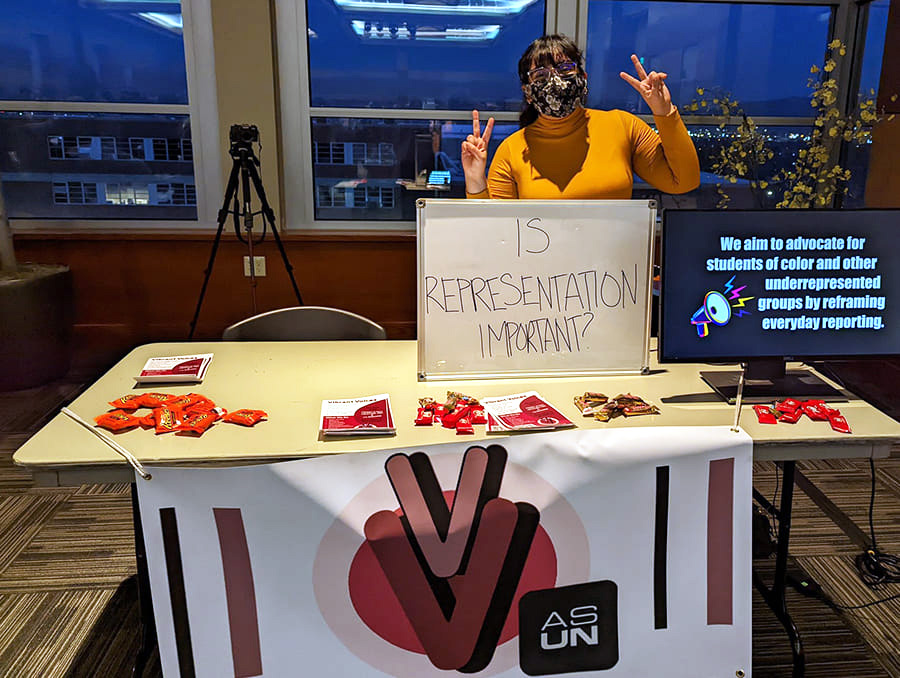 Dania Maldonado holds up peace signs with her fingers and stands behind a table with a sign on it that says "Does representation matter?"