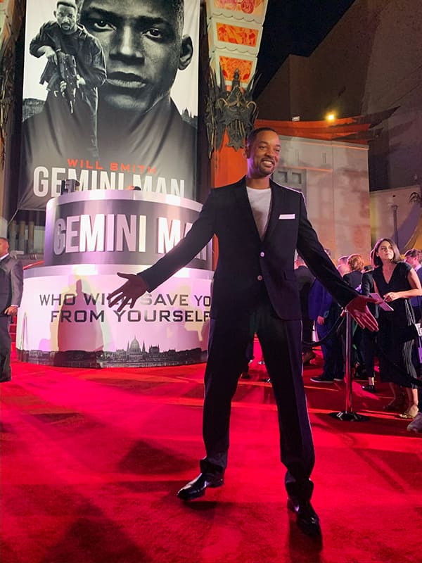 Will Smith stands on a red carpet holding his arms wide and smiling in front of a poster with a black and white picture of him and the words "Gemini Man."