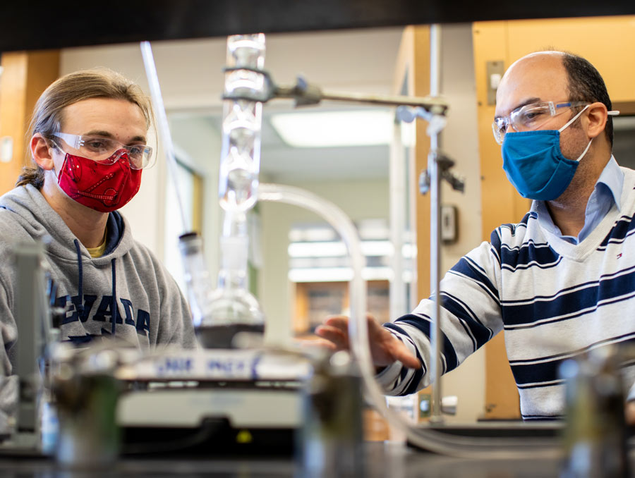 Brown (left) and Vahidi are wearing goggles and are in focus in the background, with lab equipment out of focus in the foreground.