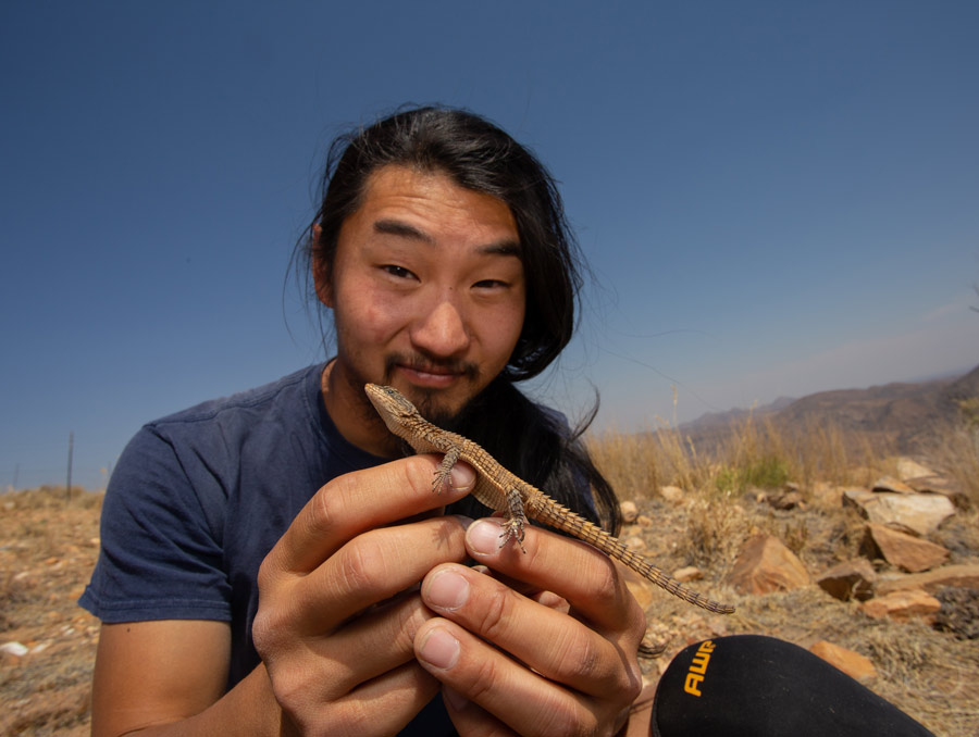 Jon DeBoer kneels, holding a brown lizard out in front of him.