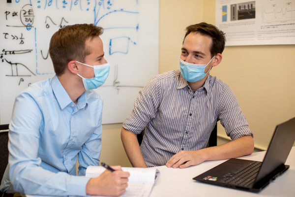 Landon Morrison (left) writes in a notebook while sitting next to Thomas White, who has a laptop in front of him. Graphs are drawn on a whiteboard in the background.