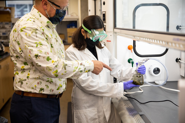 Jeffrey points to the flask being held by Stopani Barrios, who is wearing goggles and a lab coat.