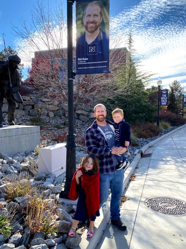 Kraus stands under his Wolf Pack Way banner, holding his son with his daughter standing next to him.