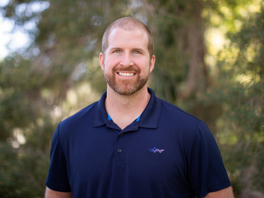 Rick Kraus smiles, wearing a blue shirt in the foreground with green foliage in the background.