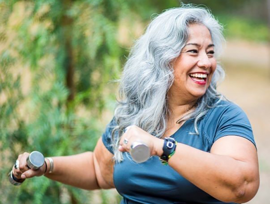 Older women with long grey hair smiling and exercising with hand weights.